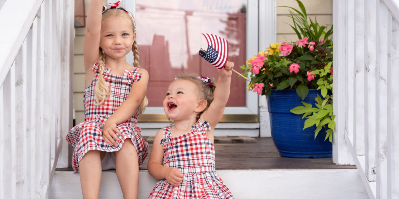 Two cute little girls sitting on the front porch on Fourth of July waving flags